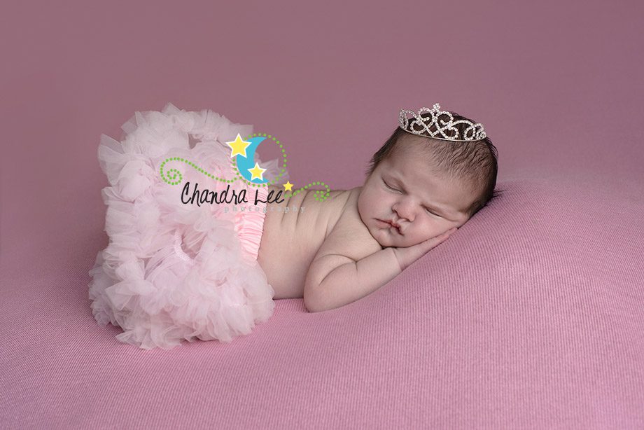 A sleeping baby with a light pink tutu and a tiara resting on a pink background.