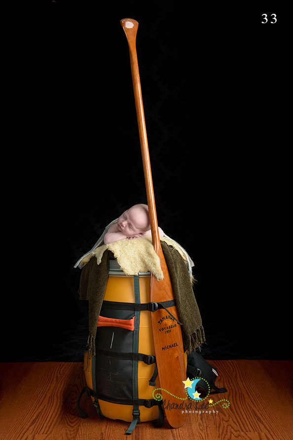 An infant asleep inside a yellow kayak, partially covered with a blanket. A wooden canoe paddle rests upright beside the baby. The background is dark, and the flooring is wooden.