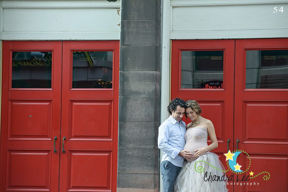 A couple stands in front of two red doors. The woman wears a strapless dress and cradles her belly, while the man in a light blue shirt stands beside her. They lean closely together, appearing intimate in a public space.