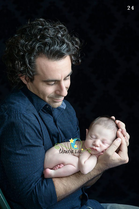 A man gently holding a sleeping newborn baby in his arms. The man is wearing a dark blue shirt and has curly hair. The background is dark with a subtle pattern.