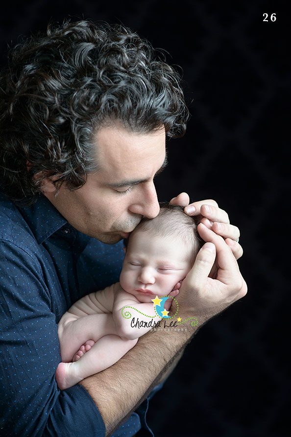 A man with curly hair gently holding and kissing a newborn baby in a studio setting. The baby is cradled in the man's hands, looking peaceful against a dark background.