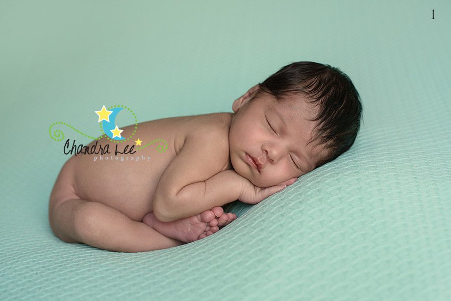 A sleeping newborn baby lying on a soft mint-green blanket, with hands under the cheek and legs curled up. The baby's hair is dark and slightly tousled.