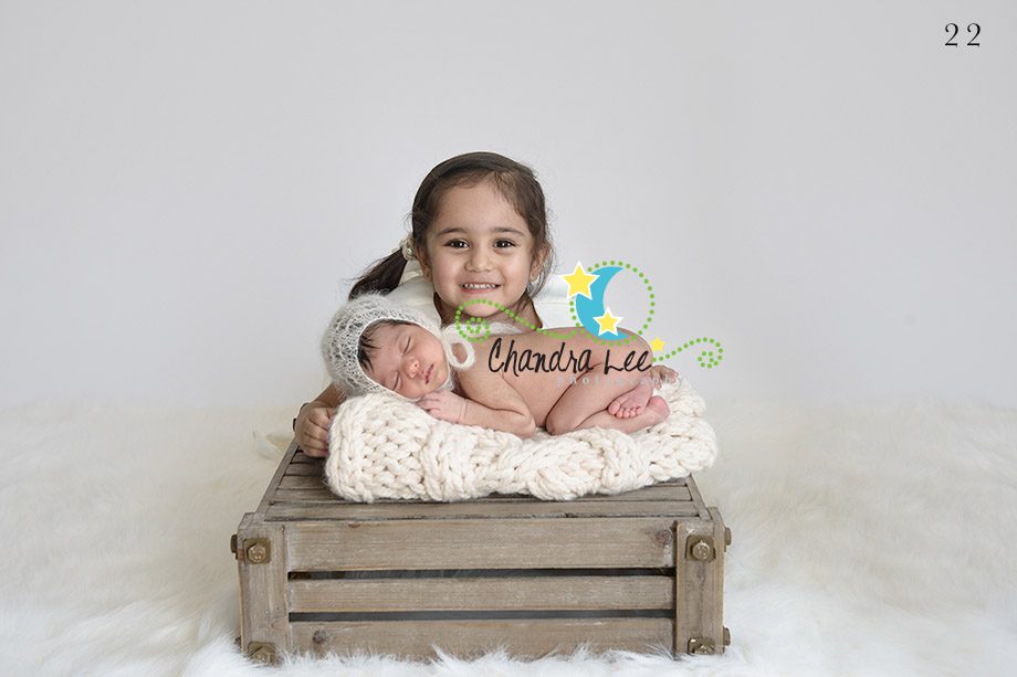 A young girl smiling while holding a wrapped newborn baby on a wooden crate. The baby is swaddled in a blanket, and both are placed on a soft, fluffy surface. The background is plain and light.