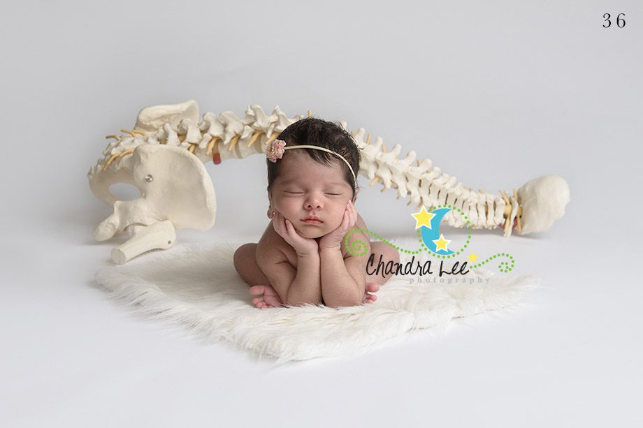 A sleeping newborn baby with hands under chin on a soft rug, beside a model of a human spine. The baby wears a small flower headband, and the background is plain white.