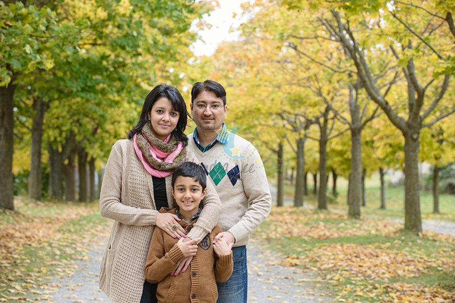 A family of three standing on a tree-lined path with autumn leaves. A woman and a man stand behind a boy, who is smiling. The woman is wearing a cream sweater and a pink scarf, while the man is in a beige sweater with patterns.