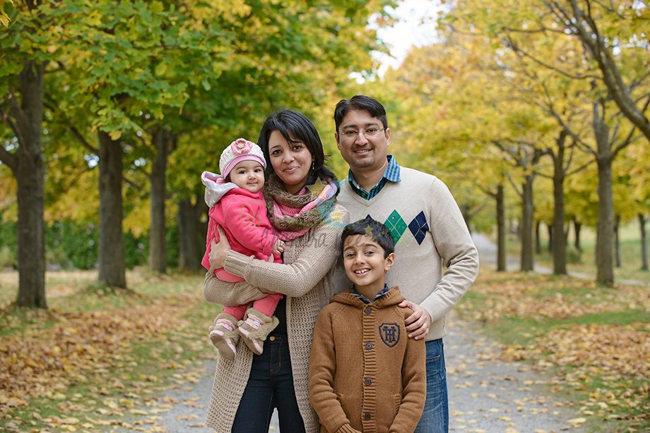 A family of four stands together on a path bordered by trees with yellow autumn leaves. The father and mother hold their two children, a baby girl in a pink jacket and a boy in a brown sweater, all smiling.