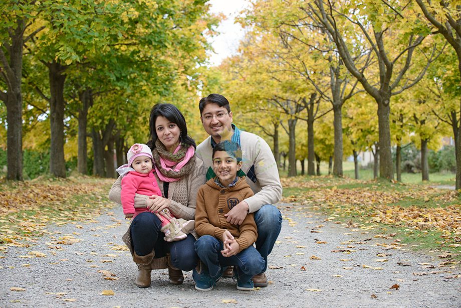 A family of four posing on a gravel path lined with trees in autumn. The mother and father are kneeling, while the mother holds a baby in a pink outfit. A young boy sits beside them, all smiling, with yellow leaves scattered on the ground.