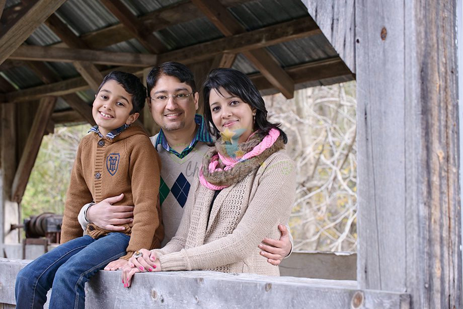 A man, woman, and child posing together at a wooden structure. The child is sitting on a railing, while the adults stand beside each other, smiling at the camera. The background shows trees and a rustic setting.