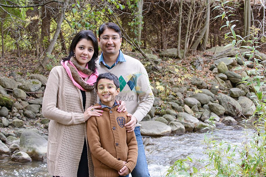 A family of three standing by a rocky stream in a forested area. The mother is wearing a light sweater and scarf, the father is in a light blue shirt and beige sweater, and the child is wearing a brown sweater. Trees are in the background.