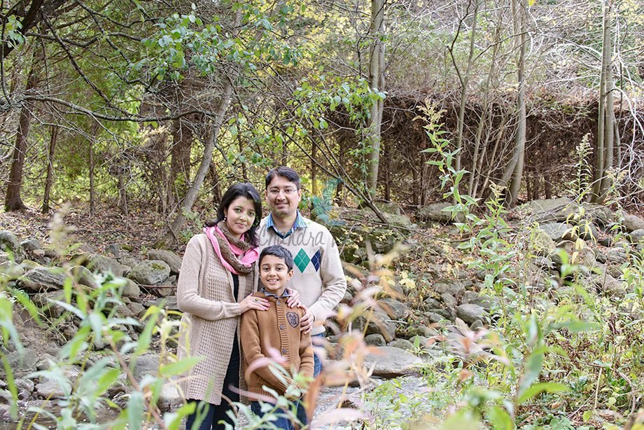 A family posing together outdoors in a natural setting. The scene shows a woman, a man, and a child standing among green foliage and rocks. They are smiling and dressed in casual, warm clothing.