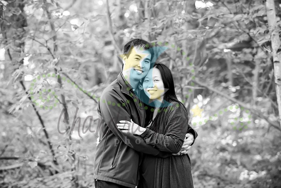 A man and a woman standing close together in a wooded area, both smiling. The background is blurred black and white, with only some color on the couple. They are wearing dark clothing and embracing each other.