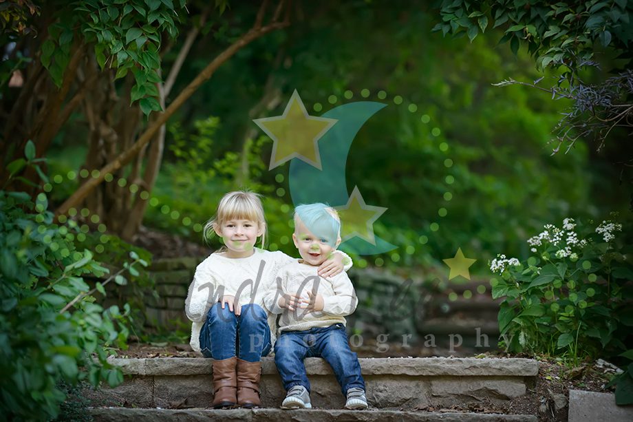 Two children sitting on stone steps in a garden. The older child has blonde hair and wears a light sweater and jeans. The younger child has blue hair, wearing a light sweater and denim pants. Green foliage and flowers surround them.