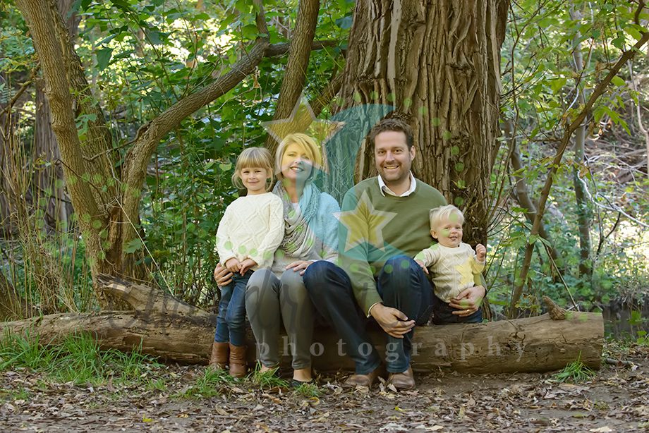 A family of four is sitting on a log in a wooded area. The family includes two children, a woman, and a man, all dressed in light-colored clothing. Green leaves and trees surround them.