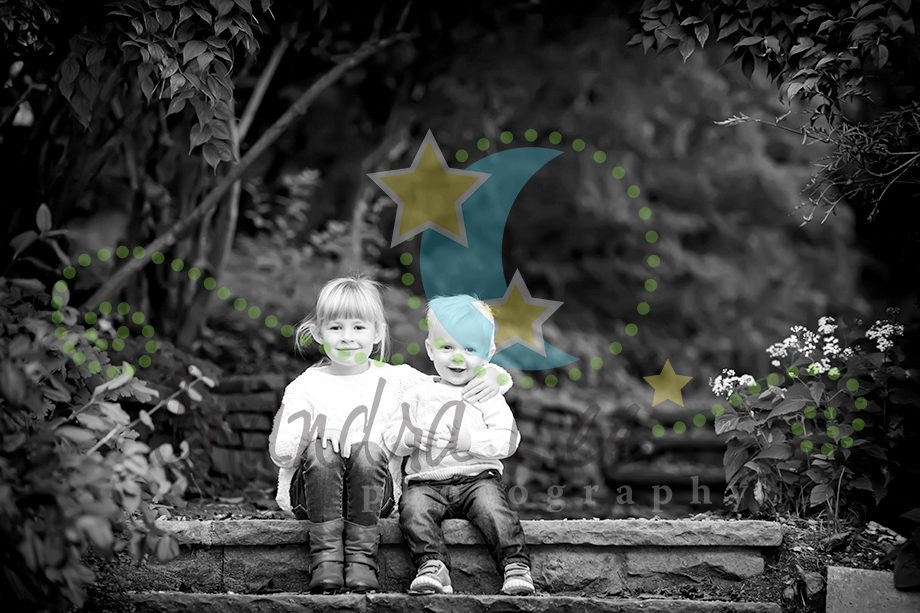 Two young children sitting on stone steps surrounded by greenery. The image is in black and white, with a watermark and graphic elements present.
