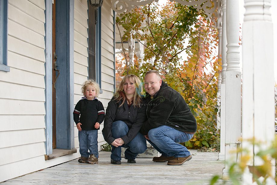 A woman, a man, and a child are posing outside a house. The child, with curly hair, is wearing a black sweater. The woman is in a gray jacket, and the man is in a black jacket. They are crouched on a wooden porch surrounded by greenery.