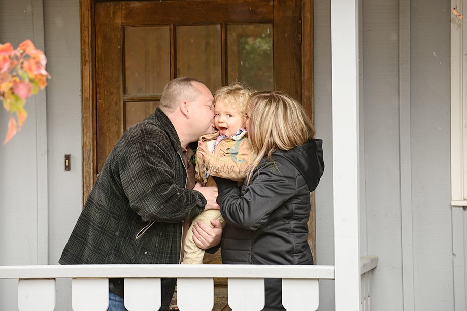 A man and a woman holding a young child outdoors on a porch. The child is smiling and looking at the camera. The adults are close to the child, with the man leaning in to kiss the child on the forehead. The setting features a wooden door and a railing.