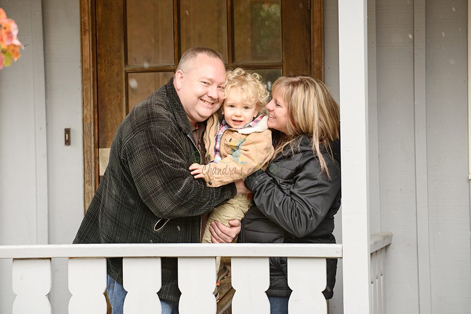 A smiling family with a man, woman, and young child, standing together on a porch. The man holds the child while the woman wraps her arm around them. The background features a wooden door and a white railing.