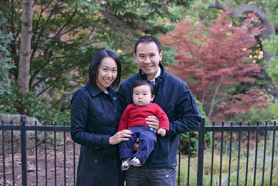 A man and a woman pose outdoors for a photo with a baby in the woman's arms. The woman wears a dark coat, and the baby is dressed in a red sweater. Colorful foliage and a fence are visible in the background.