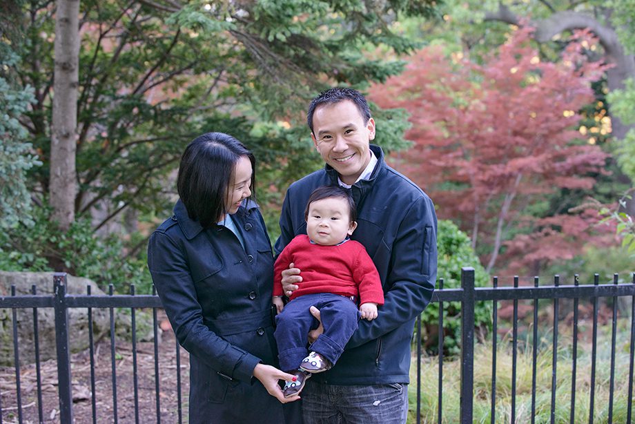 A man and a woman standing outdoors, holding a baby. The baby is wearing a red sweater and smiling. The background features greenery and a wooden fence.