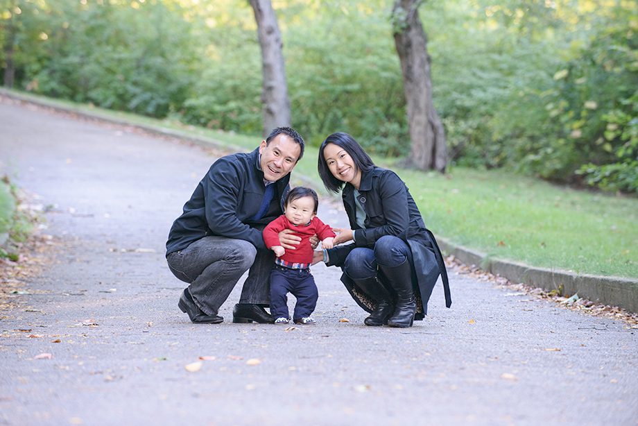 A man and a woman crouching beside a small child on a pathway in a park. The child is wearing a red sweater and dark pants. The background features green foliage and trees.