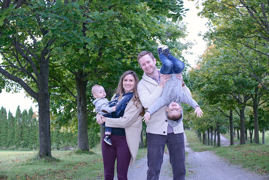 A family of four standing on a gravel path among trees. The mother holds a baby, while the father holds a boy upside down. The other boy is balancing on the father's shoulder. They are all smiling.