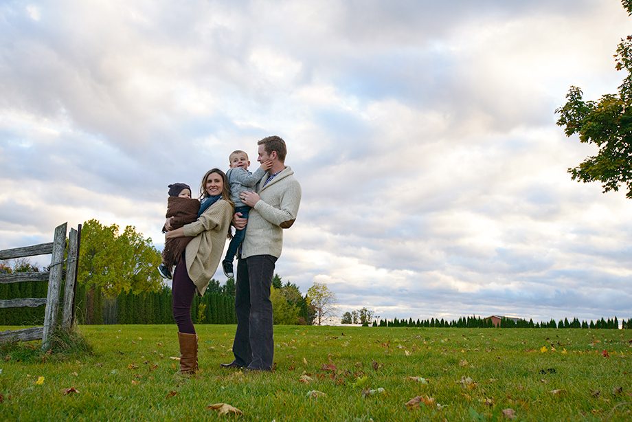 A family standing in a grassy field. The mother holds a baby and smiles, while the father carries a child on his shoulder. A dog is at the mother's feet. There are trees in the background and fluffy clouds in the sky.