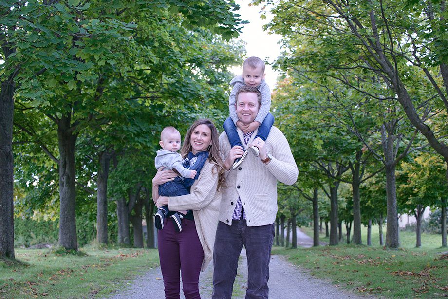 A family of four stands on a gravel path lined with trees. The father is holding a toddler on his shoulders, while the mother carries an infant in her arms. They are dressed casually, with smiles visible as they pose together.