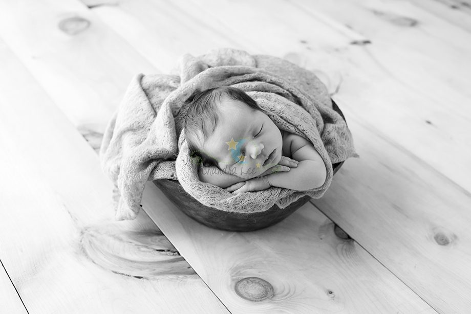 A newborn baby wrapped in a gray blanket is sleeping inside a wooden bowl, placed on a light-colored wooden floor. The image is in black and white.