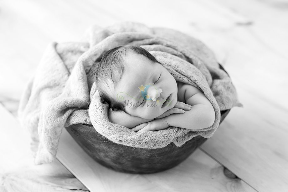 A newborn baby peacefully sleeping on a soft blanket inside a wooden bowl. The image is in black and white, showcasing the baby's relaxed posture and cute features.