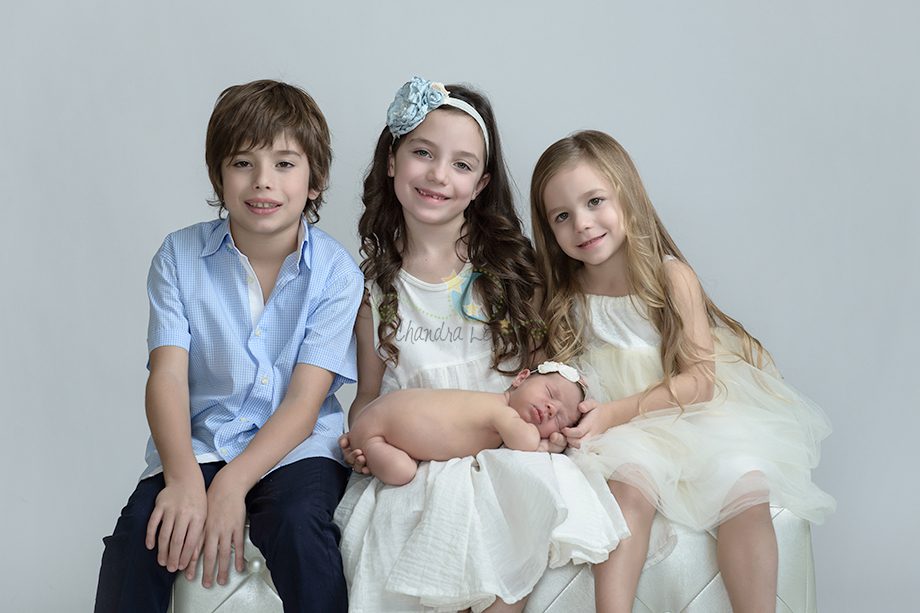 Three children pose for a photo against a light gray background. The boy on the left wears a blue shirt, the girl in the middle has a headband and smiles, and the girl on the right wears a white dress. An infant is cradled in the girl's lap.
