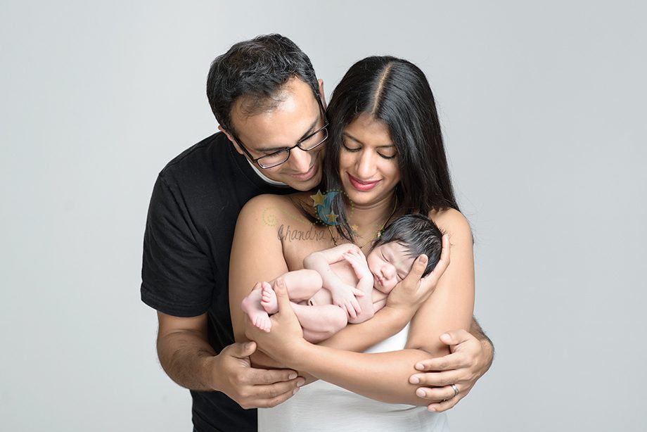 A couple holding a newborn baby between them, with the father in a black t-shirt and the mother in a white top. They are standing in front of a light gray background, both smiling down at the baby.