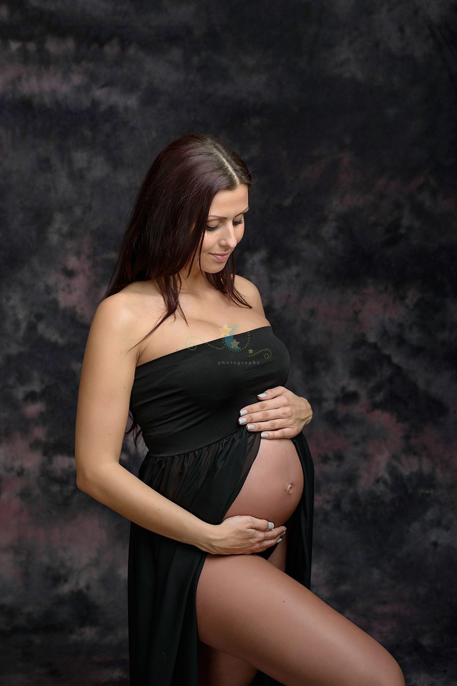 A pregnant woman in a black strapless dress standing in a studio. She is gently holding her belly with one hand while looking down, against a blurred dark background.