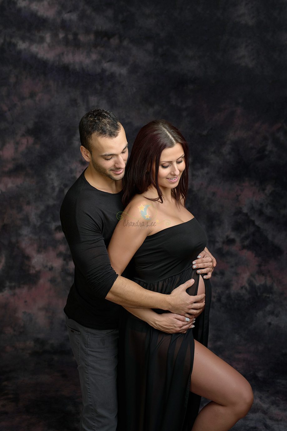 A couple standing closely together, with a man gently embracing a woman who is pregnant. The woman wears a black dress that drapes over her belly. The background is a textured blend of dark colors.