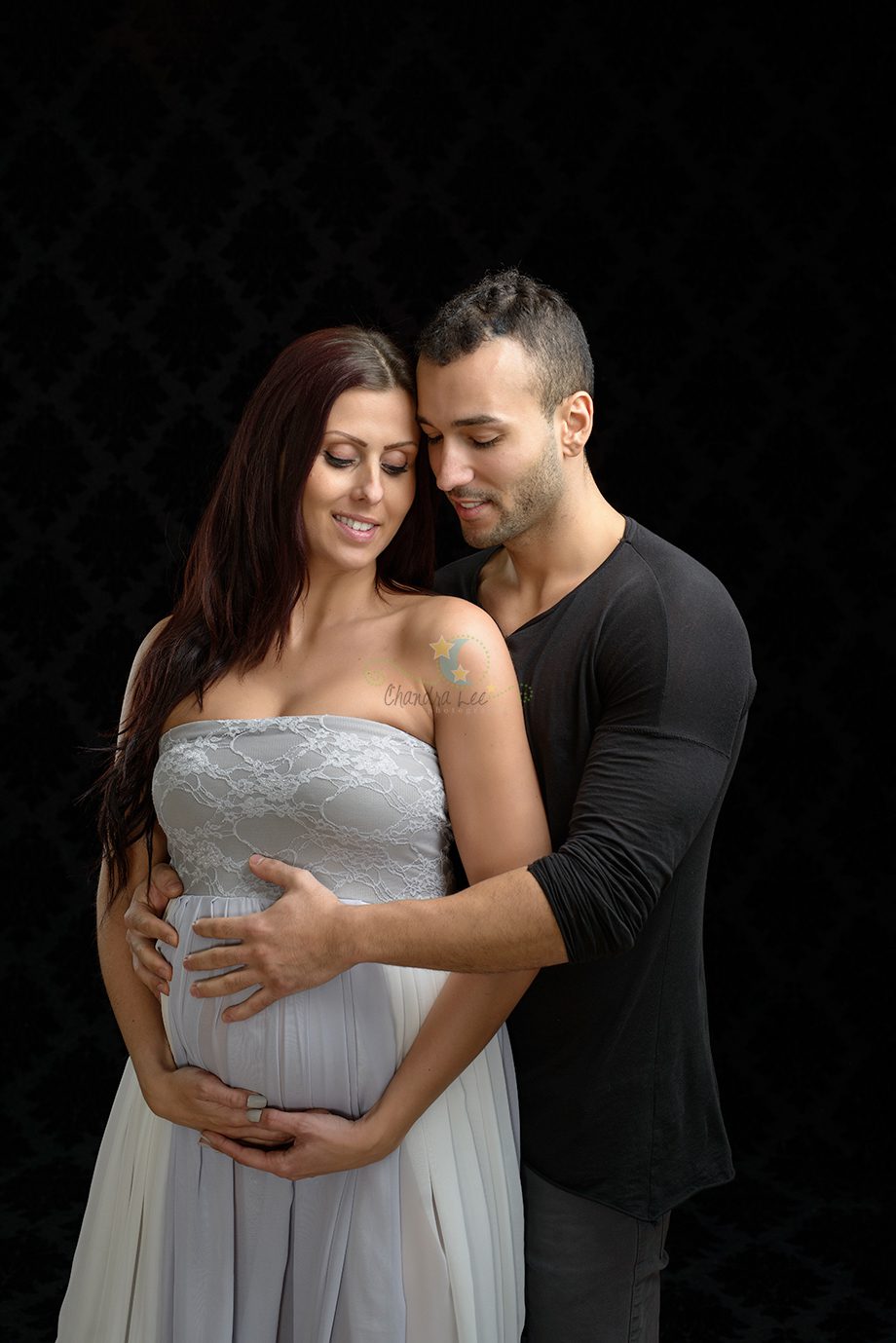 A pregnant woman in a light-colored dress is standing next to a man wearing a black shirt. The man is gently resting his hands on the woman's stomach. The background is dark, highlighting the couple's pose.