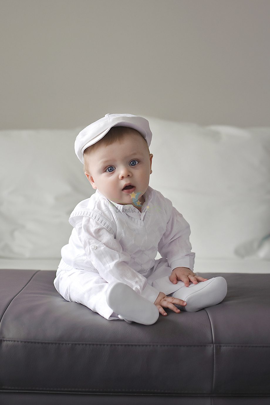 A baby sitting on a gray couch wearing a white outfit and a matching cap, looking towards the camera. The background is soft-focus with a light-colored sofa.