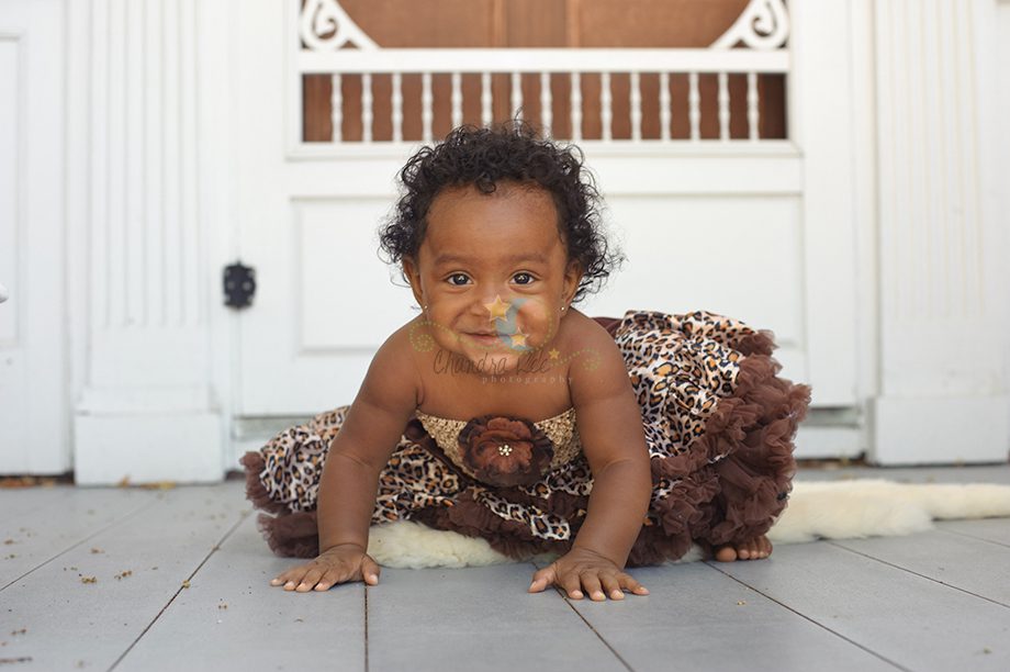 An infant crawling on a wooden floor, wearing a brown and leopard print dress with a flower detail. The background features a white wall and a doorway.