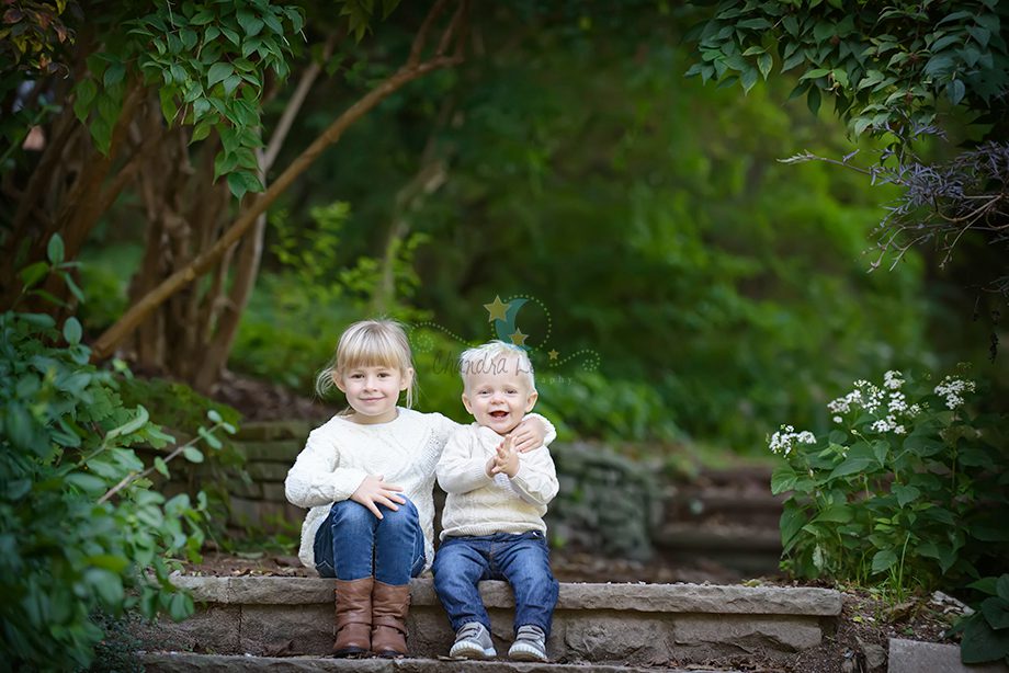 A girl and a boy sitting on stone steps in a garden. The girl wears a white sweater and blue jeans with brown boots. The boy wears a light sweater and blue pants. Green foliage and flowers surround them.