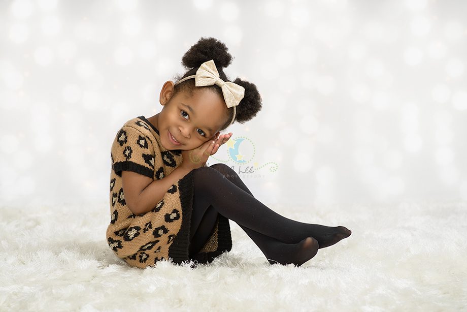 A young girl with curly hair styled in buns, wearing a brown leopard-print dress and black tights, sits on a fluffy white surface with a playful expression. The background has a soft, blurred design with bright spots.