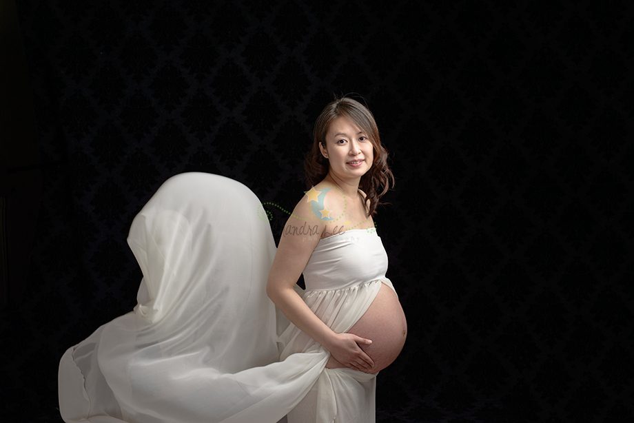 A pregnant woman in a white dress stands against a dark background. She is smiling and holds her belly with one hand. A sheer white fabric flows behind her, creating a soft, flowing effect.