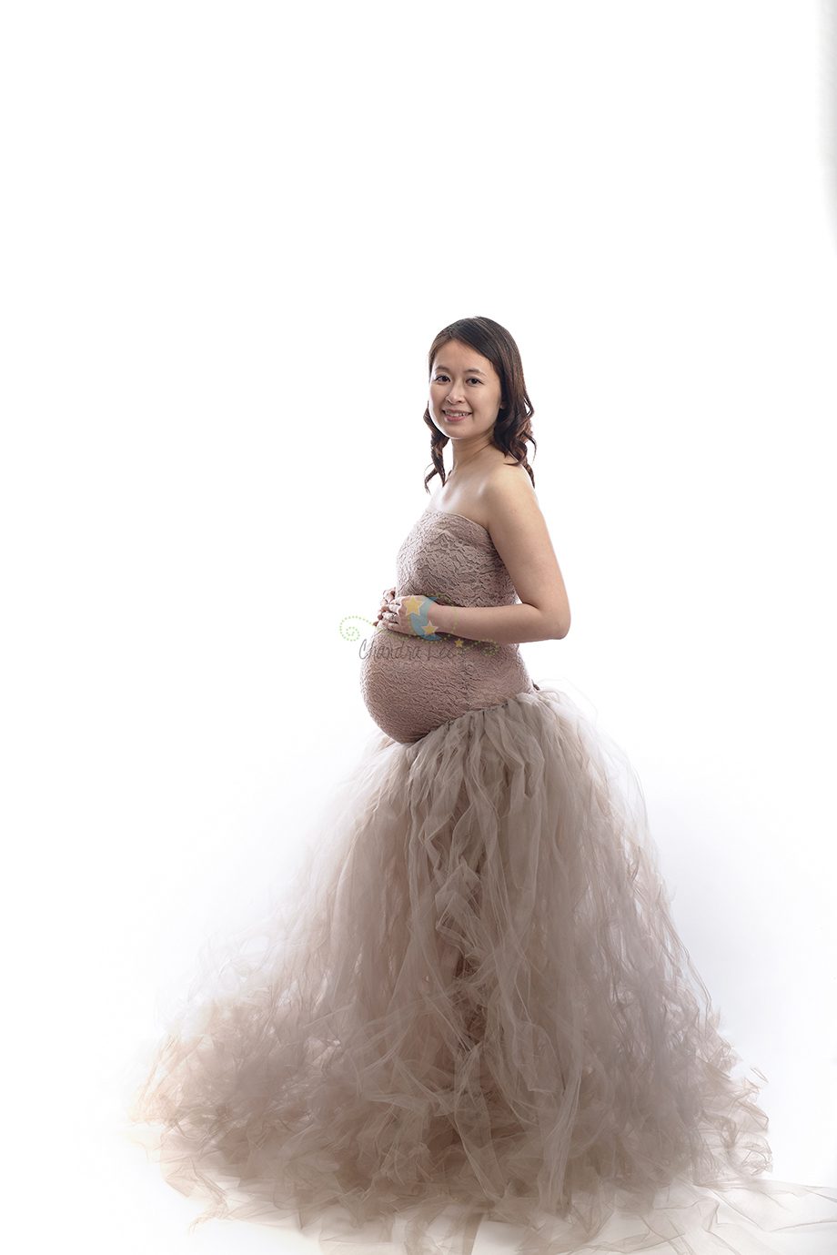 A pregnant woman wearing a strapless, form-fitting dress with a tulle skirt, standing against a white background. She is holding a baby item with both hands and smiling at the camera.