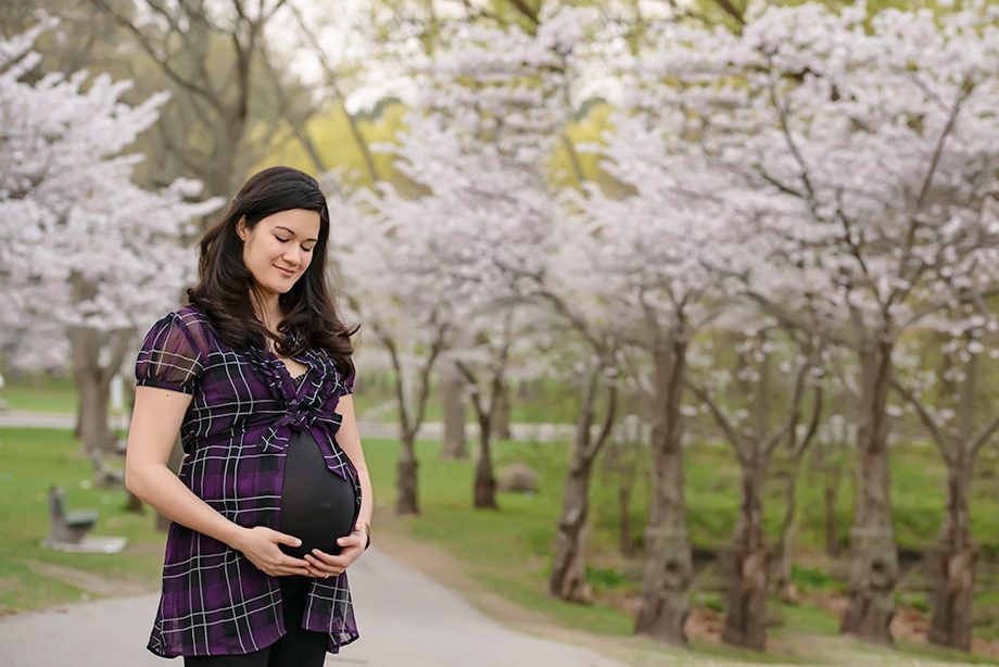 A woman with long dark hair stands outdoors, gently holding her pregnant belly. She is wearing a purple plaid top and black pants, with cherry blossom trees blooming in the background along a pathway.