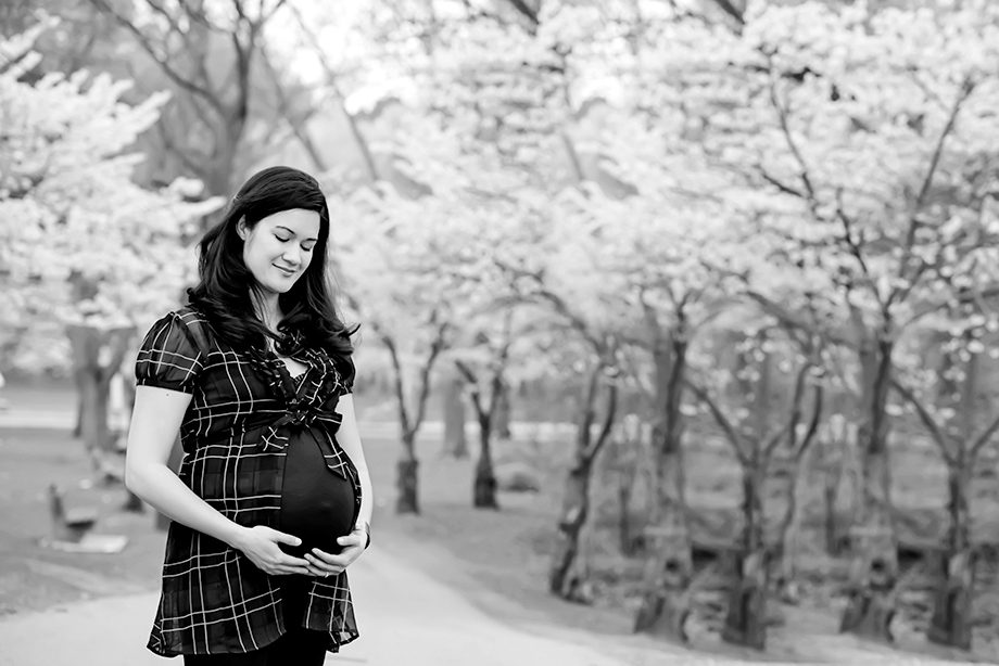 A pregnant woman in a black and white setting is standing outdoors, smiling while holding her belly. She is wearing a black dress with a checkered pattern, surrounded by blooming trees in the background.