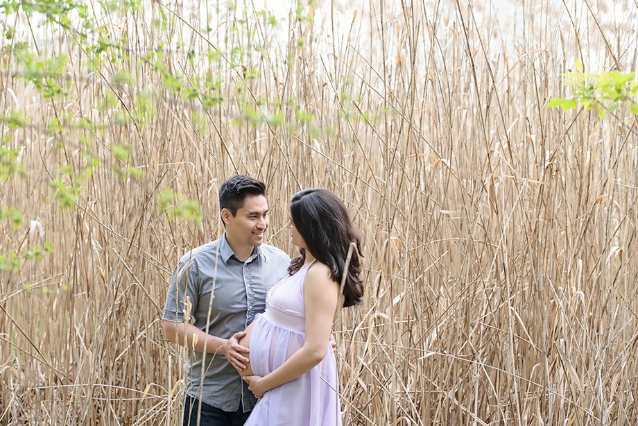 A couple stands in a field of tall, dry grass. The man wears a gray shirt, and the woman, showing her pregnancy, wears a light purple dress. They are smiling at each other, surrounded by greenery.
