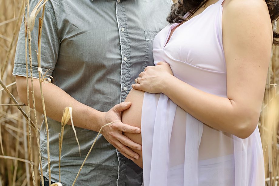 A pregnant woman in a flowing white dress holds her belly, while a man stands beside her with one hand on her waist in a field of tall grass.