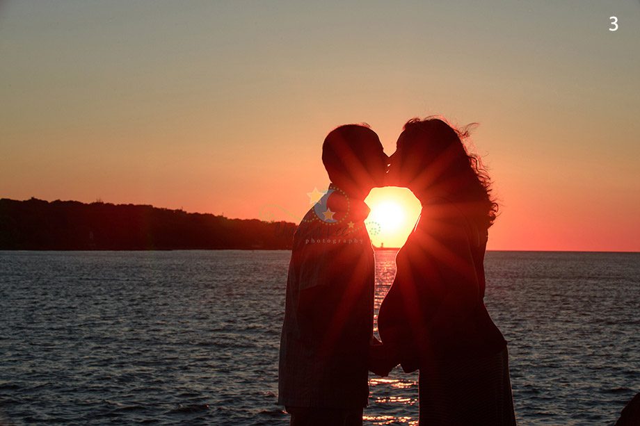 A couple facing each other and kissing with a sunset in the background over a body of water. Silhouette of the couple is visible against the colorful sky.