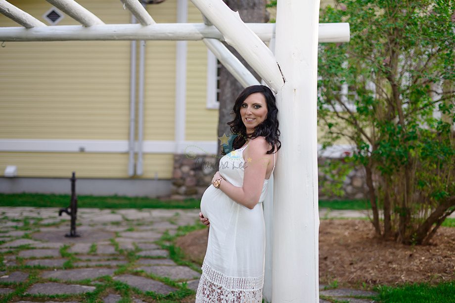 A woman with long dark hair wearing a white dress stands outside, leaning against a white wooden structure. She is showing a visible baby bump and smiling. The background includes green grass and trees.