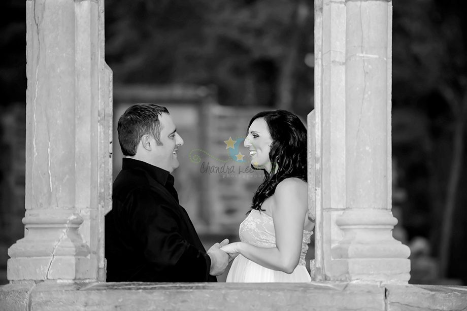 A couple stands in a stone archway, facing each other, holding hands. The man is wearing a black shirt, and the woman is in a white dress. The background is slightly blurred, revealing trees and a stone structure.