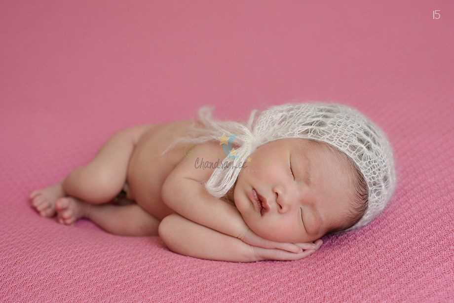A newborn baby lying on a pink background, wearing a delicate white knitted bonnet. The baby is sleeping peacefully with hands tucked under the chin.