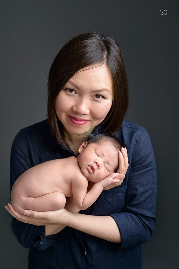 A woman holding a newborn baby cradled in her hands. The baby is asleep and naked, while the woman is smiling and wearing a dark blue shirt, set against a gray background.