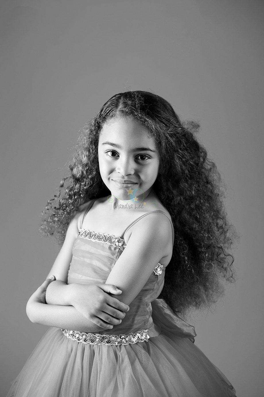 A young girl with long, curly hair, wearing a sleeveless dress with a decorative band, stands with her arms crossed. The image is in black and white with a simple background.